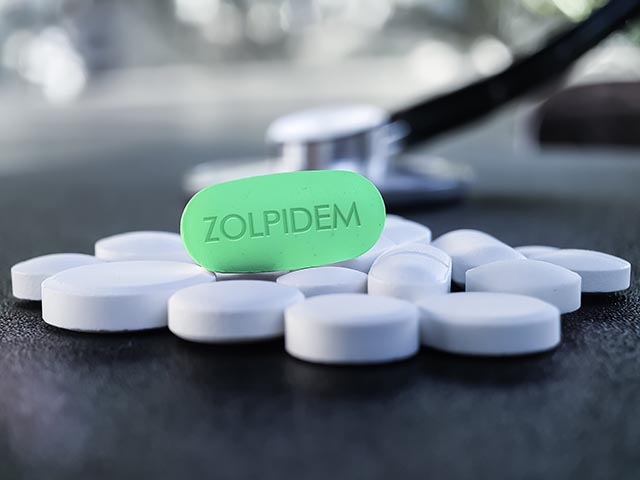 Ambien pills sit on a table in front of a stethoscope, with a large pill saying Zolpidem, the brand name for Ambien, sits on top. 