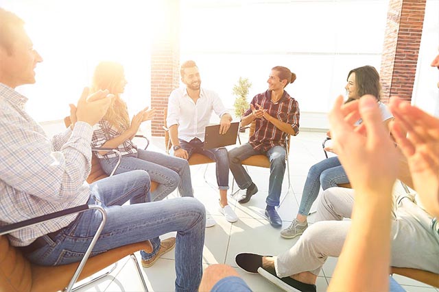 A group of people in outpatient treatment for addictions (including heroin) smile during a process group with a therapist and peers.