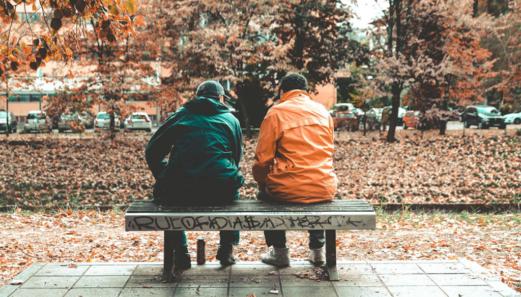 A man helps another to recover from his addiction as they sit on a park bench talking - the importance of giving back.