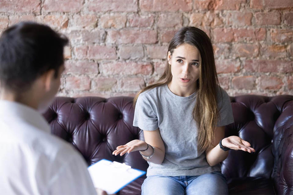 A woman shrugs innocently to a therapist or counselor, claiming she does not have a problem with drugs or alcohol.