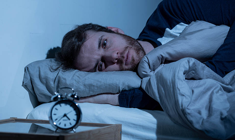 A restless man lies awake at night staring at the clock while detoxing from alcohol.