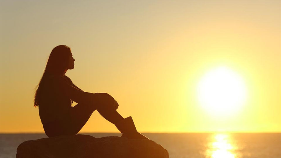 A woman relaxes by the ocean during sunset, reducing stress to help alleviate drug and alcohol cravings.