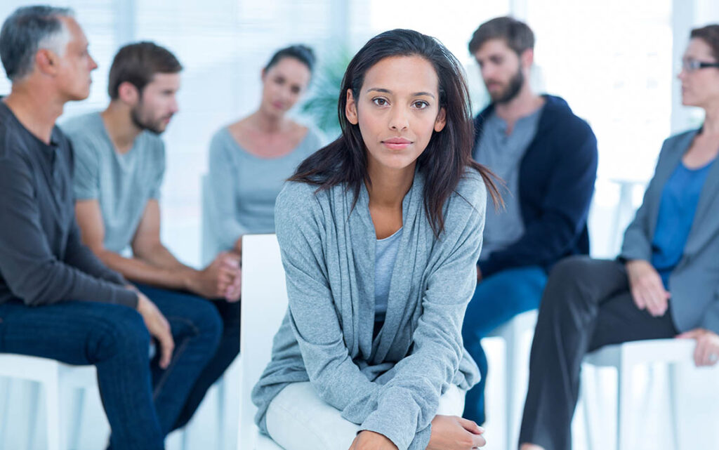 A woman sits looking at the camera while in the blurred background an outpatient addiction group takes place.