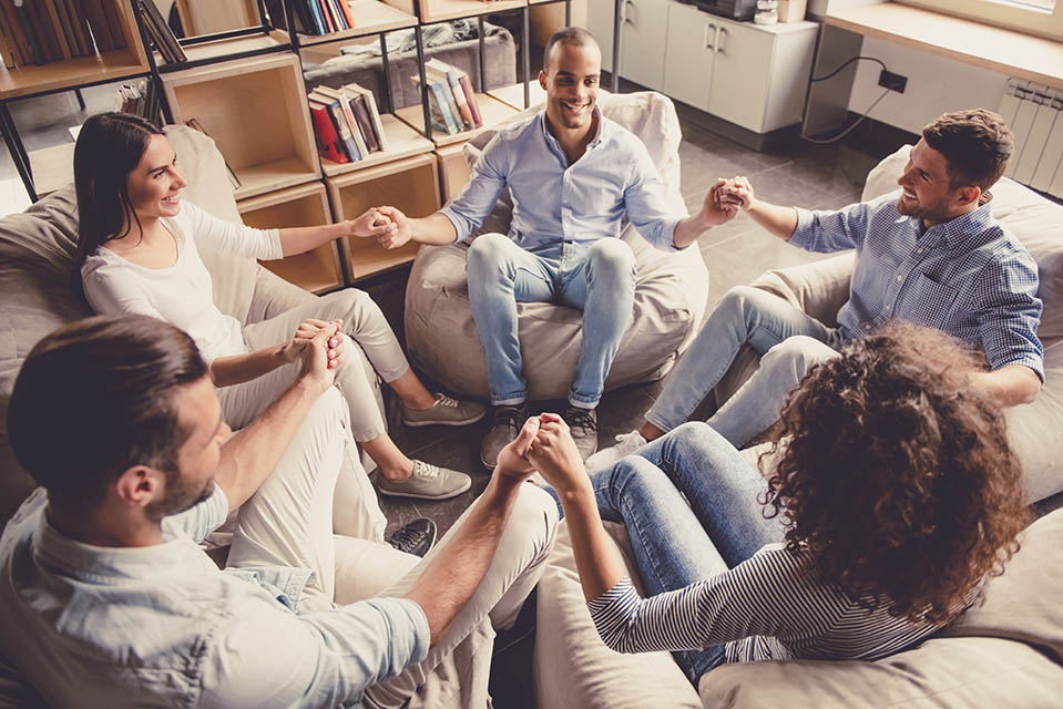 Young people are sitting in circle, holding hands and smiling at an out-of-state rehab center for addiction.