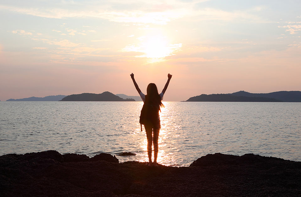 A woman stands by the beach celebrating with her arms in the air, feeling good after getting sober and staying away from old triggers