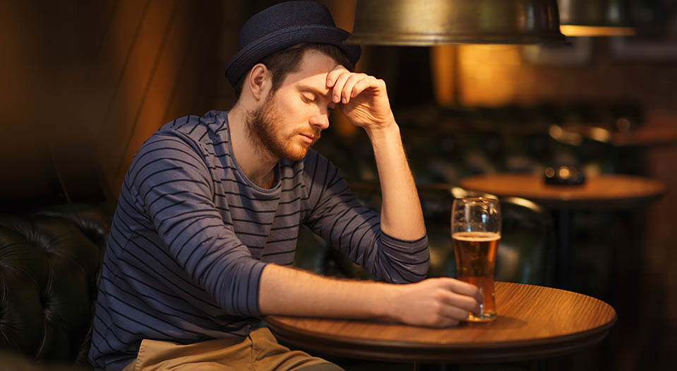 A man sits alone at a table looking sad and drinking, showing the link between alcohol and mental health.