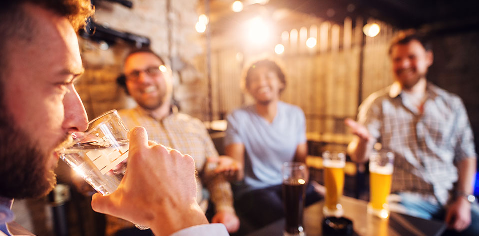 A group of guys poke fun at their friend for drinking water, showing being sober when others are drinking alcohol is a common and stressful addiction trigger.