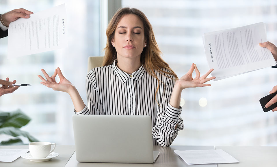 An over-worked woman dealing with a lot of stress takes a break to meditate while coworkers hold out work papers - symbolizing tips for dealing with stress.