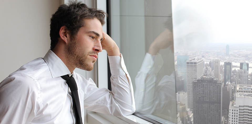 A stressed office worker looks out the window as his reflection shows, symbolizing self reflection.