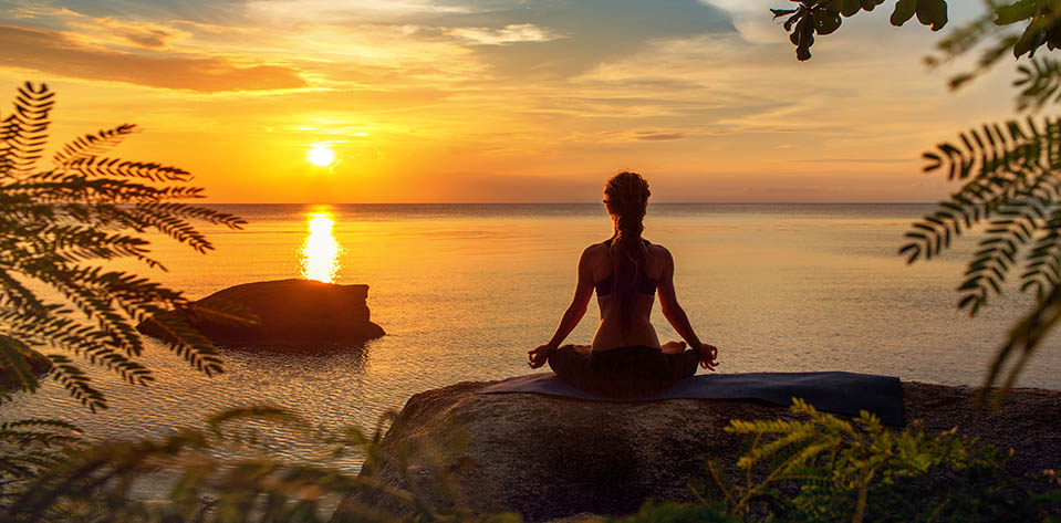 A lady experiencing stress takes times to meditate on the shore near the water at sunset.