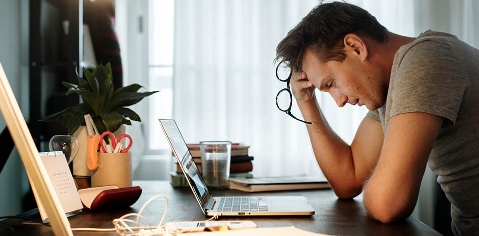 An overworked man sits in front of his computer with his glasses and head in his hand, attempting to get in touch with his stress. Becoming a student of stress.