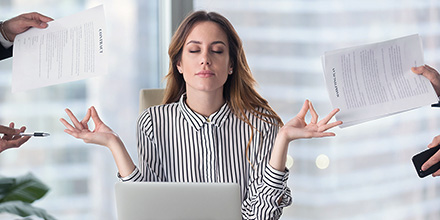 An over-worked woman dealing with a lot of stress takes a break to meditate while coworkers hold out work papers - symbolizing tips for dealing with stress.