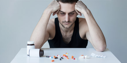 Man struggling with co-occurring addiction and mental health problems looks sad with hands on head, resting elbows on table as he stares at a spilled pill bottle of drugs.