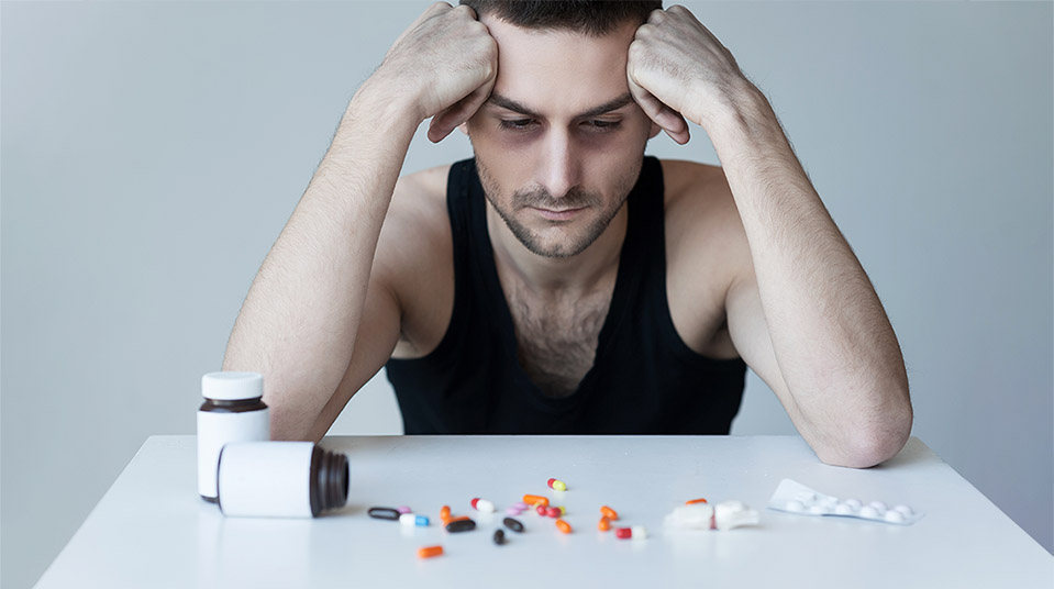 Man struggling with co-occurring addiction and mental health problems looks sad with hands on head, resting elbows on table as he stares at a spilled pill bottle of drugs.