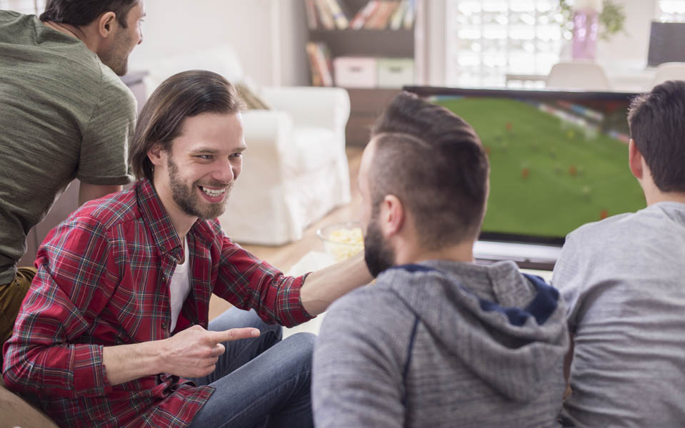 Two friends laugh together as a group of guys watch TV together while living together as roommates in Sober Living.