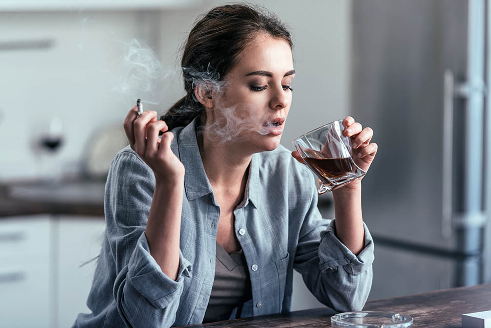 A woman struggling with co-occurring substance abuse and clinical depression drinks alcohol and smokes a cigarette in her home.