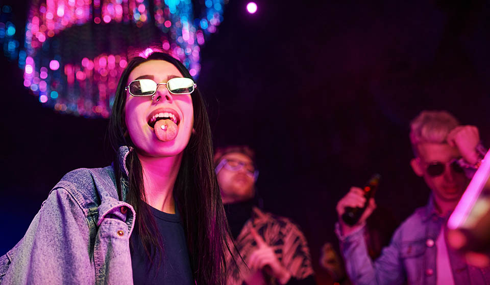 A young woman experimenting with drugs sticks her tongue out, showing the pill she is about to take at a nightclub.