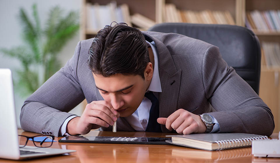 A well groomed young man wearing a suit in the second or third stage of addiction leans forward to snort drugs off a desk in his office at work.