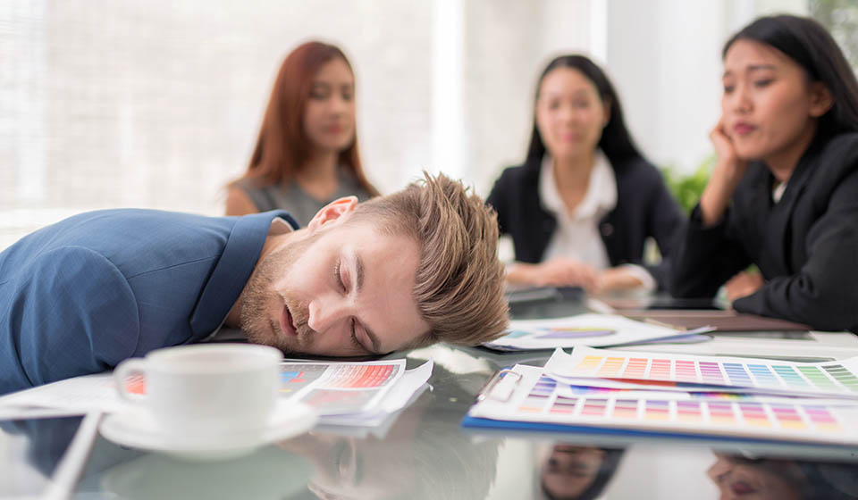 A man in the high-risk phase of addiction passes out with his face on the table during a business meeting, high from using drugs while unamused coworkers look at him.