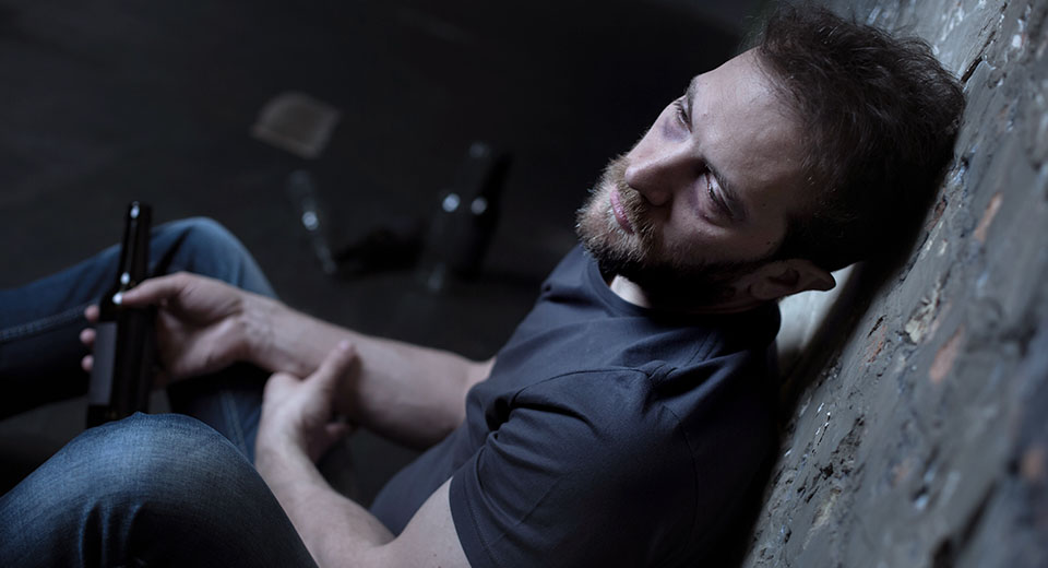 An alcoholic deep in his addiction sits with his back against a broken wall in the dark with a beer bottle in his hand looking worn down and depressed.