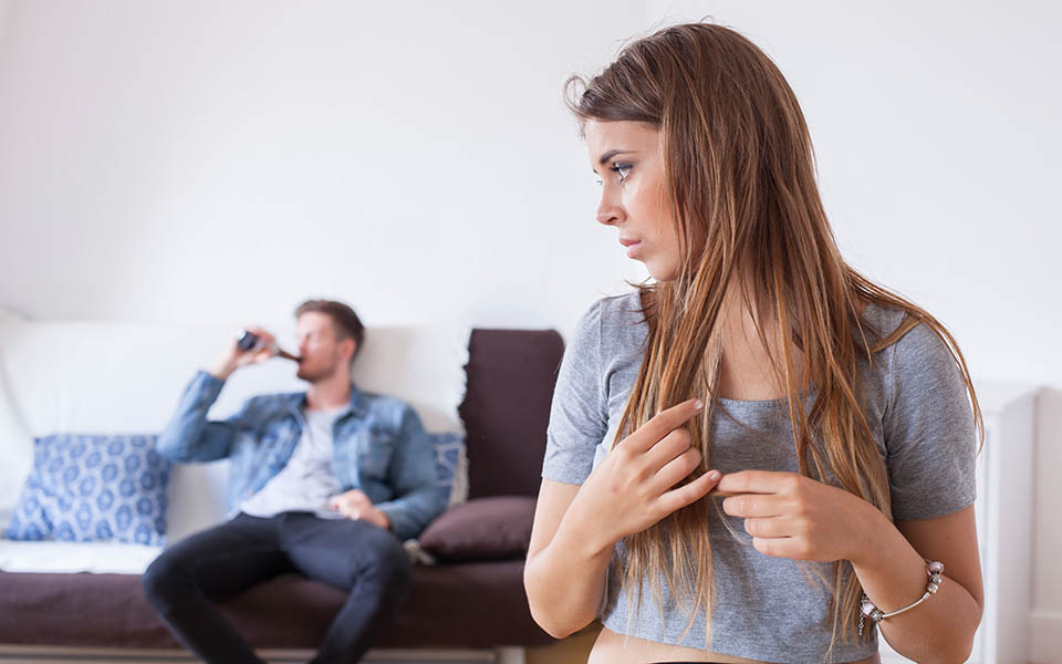 An upset woman contemplates how to help her husband who is addicted to alcohol and drinking in the background.