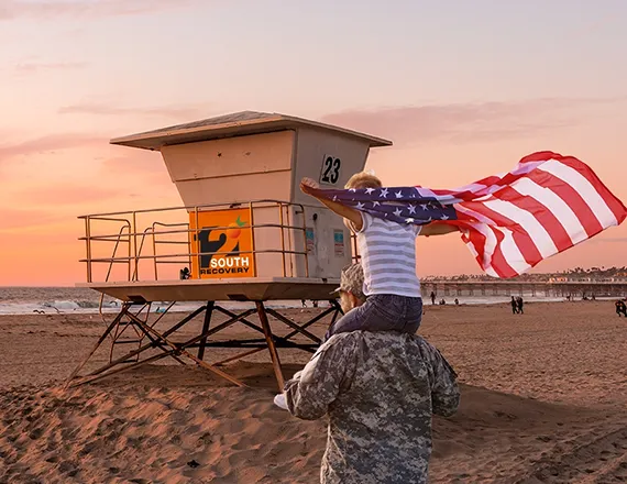 A veteran receiving help walks in front of a lifeguard tower that is painted with the 12 South Recovery logo, with his child on his shoulders wearing an American flag as a cape, showing that 12 South Recovery specializes in mental health and addiction treatment for veterans.