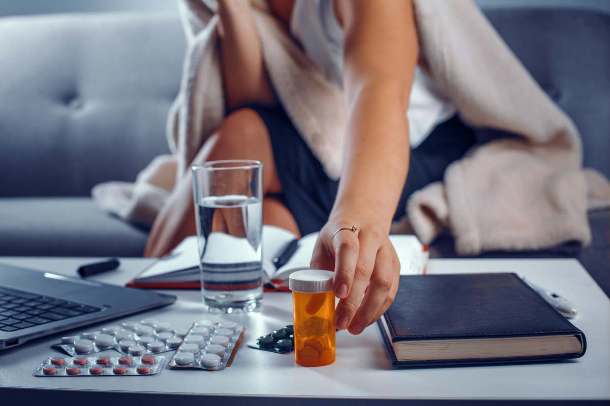 Woman's hand with a ring on an index finger reaches for the pills in a container. Notebook, laptop, glass of water and other medicines in tablet packs are laying on the table.