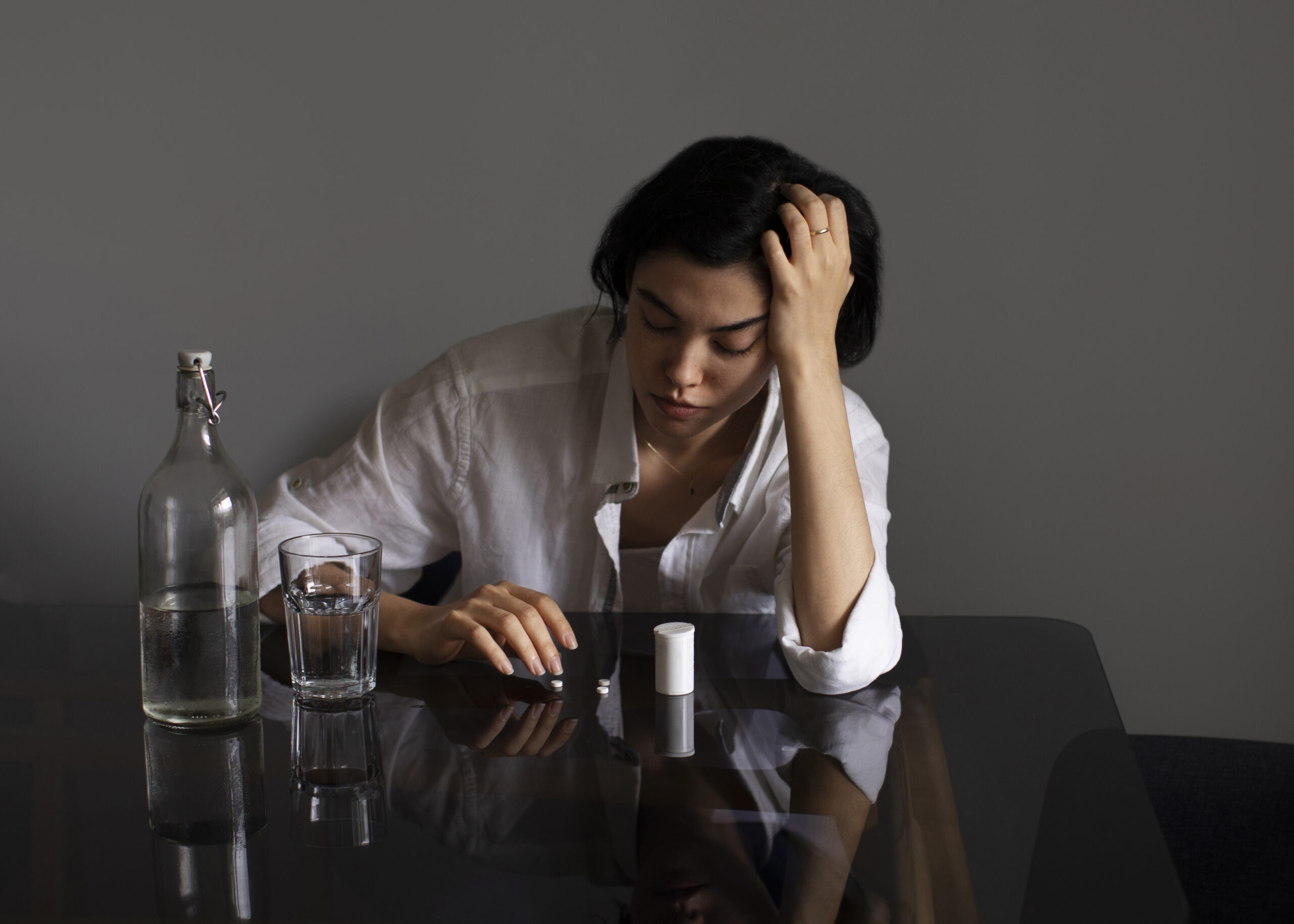 Woman hungover leaning on a table stressed with a glass of water and pills