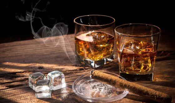 Two glasses of whiskey with ice cubes, a lit cigar on an ashtray, and extra ice on a rustic wooden table, with rising smoke in a dimly lit setting.