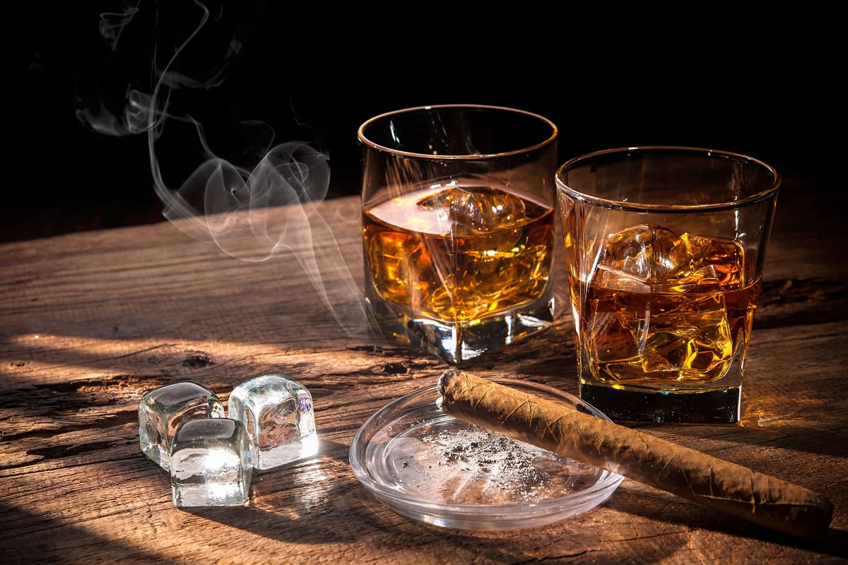 Two glasses of whiskey with ice cubes, a lit cigar on an ashtray, and extra ice on a rustic wooden table, with rising smoke in a dimly lit setting.