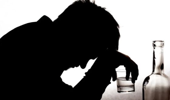 Silhouette of a person holding a glass and leaning over in distress beside a bottle, symbolizing emotional struggles and potential addiction to dark liquor.