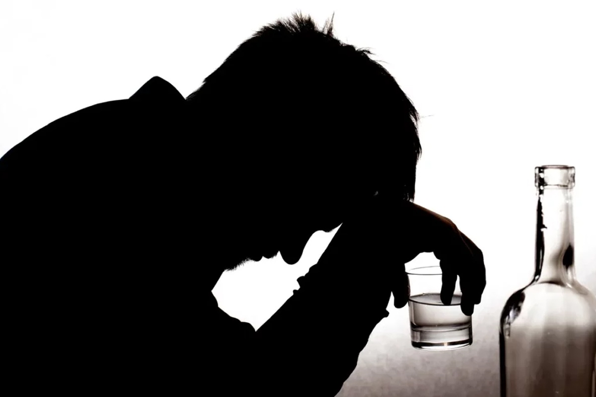 Silhouette of a person holding a glass and leaning over in distress beside a bottle, symbolizing emotional struggles and potential addiction to dark liquor.